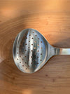 A stainless steel slotted spoon with a perforated bowl, shown on a warm-toned wooden surface.