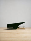 A modern, minimalist desk staple in a dark forest green finish with a polished gold-toned metal hinge, resting on a light wood surface against a neutral light gray background.