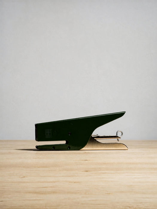 A modern, minimalist desk staple in a dark forest green finish with a polished gold-toned metal hinge, resting on a light wood surface against a neutral light gray background.