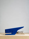 A blue metal stapler with silver metal components, positioned on a light wood surface against a neutral light gray background.