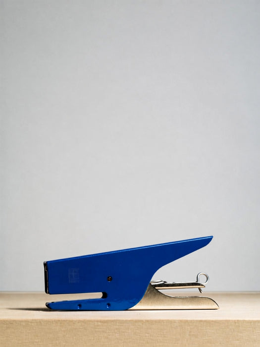 A blue metal stapler with silver metal components, positioned on a light wood surface against a neutral light gray background.