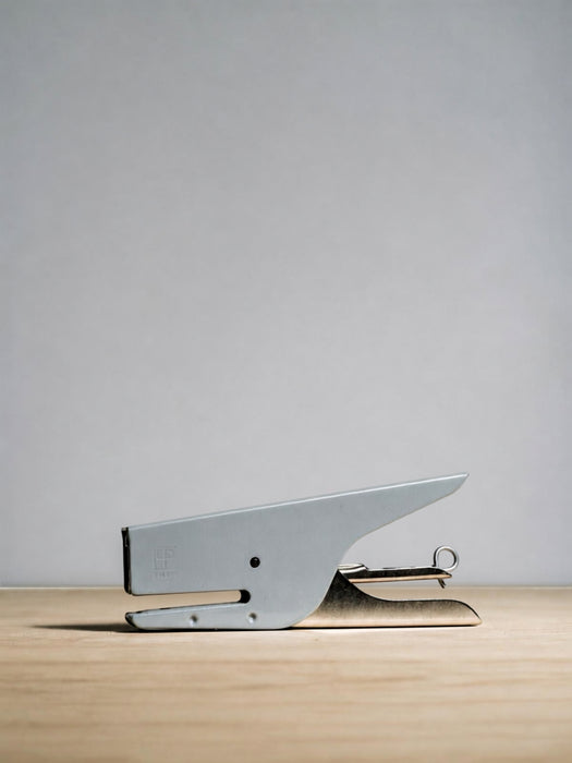 A silver metal stapler resting on a light wood surface against a neutral gray background.