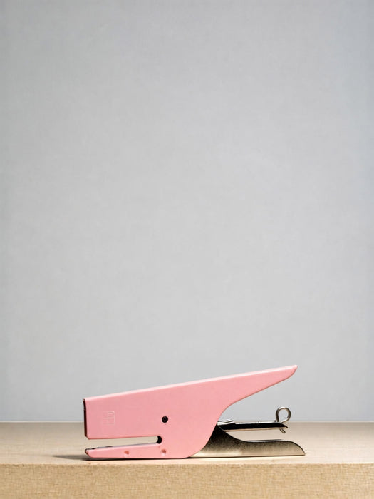A pink metal stapler with a silver metal hinge, resting on a light beige wooden surface against a solid light gray background.