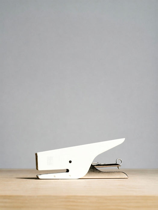A minimalist white desk staple with a sleek, modern design resting on a light wood surface against a solid gray background.