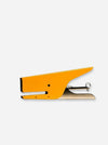 A bright yellow-orange metal staple with a silver-toned metal spring and a small black rubber tip, shown on a plain light gray background.