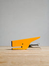 A bright yellow desk stapler with a silver metal hinge and staple mechanism, resting on a light wood surface against a neutral gray background.