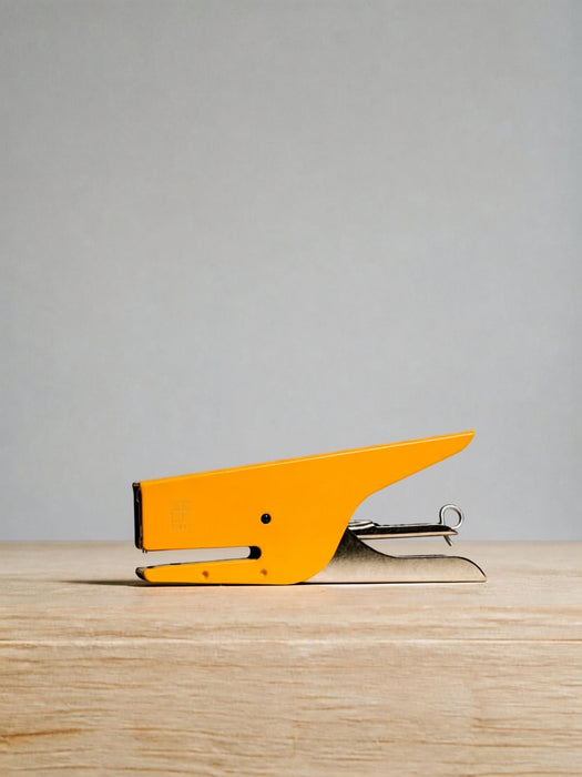 A bright yellow desk stapler with a silver metal hinge and staple mechanism, resting on a light wood surface against a neutral gray background.