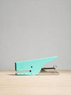 A small turquoise metal paperclip with a silver spring and guide bars, resting on a light wood surface against a neutral gray background.
