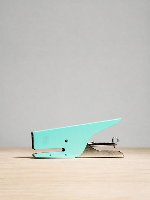 A small turquoise metal paperclip with a silver spring and guide bars, resting on a light wood surface against a neutral gray background.