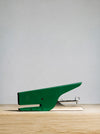 A green and silver stapler placed on a light wood surface against a neutral gray background.
