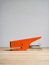 A bright orange metal stapler with a silver metal hinge and staple mechanism, resting on a light wood surface against a neutral gray background.