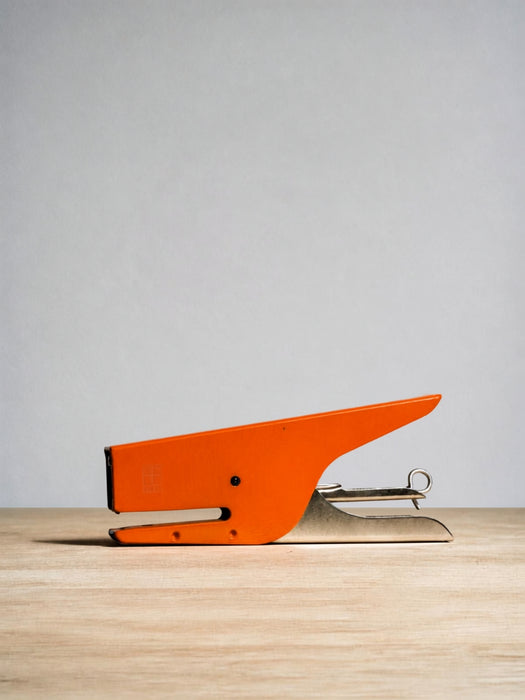 A bright orange metal stapler with a silver metal hinge and staple mechanism, resting on a light wood surface against a neutral gray background.