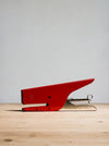 A red metal stapler with silver metal blades and hinge, resting on a light-colored wooden surface against a neutral gray background.