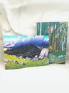 Two square art prints or cards showing painted landscape scenes, one with a mountain and wildflower meadow, and one with a forest scene, displayed against a white fabric backdrop.