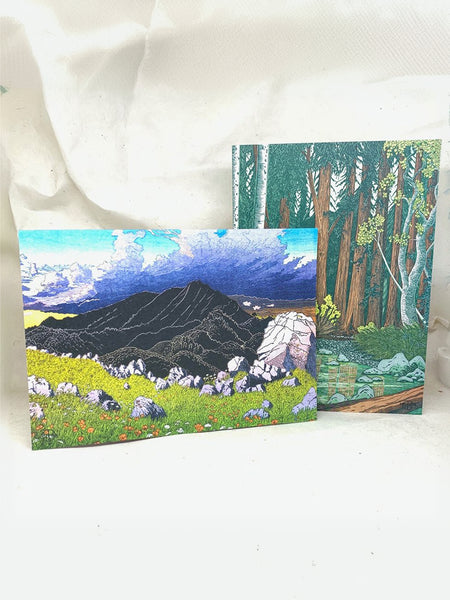 Two square art prints or cards showing painted landscape scenes, one with a mountain and wildflower meadow, and one with a forest scene, displayed against a white fabric backdrop.