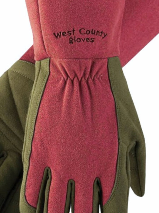 A close-up of a heavy-duty work glove with a dusty rose pink leather palm and fingers, olive green leather back and wrist, and black embroidered text reading 'West County gloves'.