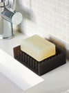 A rectangular translucent soap bar resting on a dark brown silicone soap dish with drainage slots, placed on a white bathroom counter next to a chrome faucet and tiled wall.