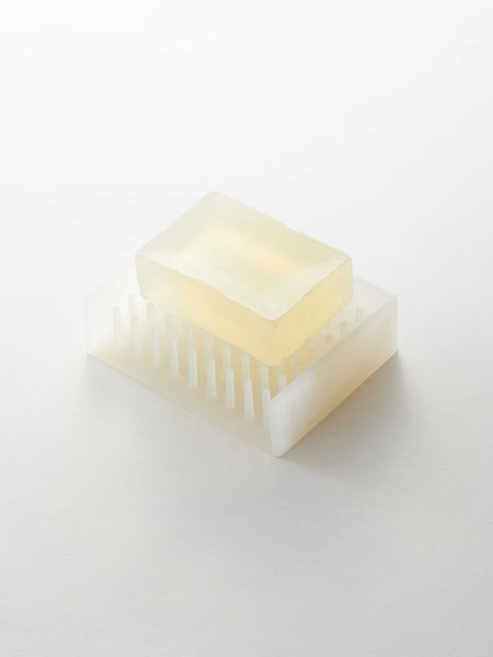 A translucent pale yellow rectangular bar of soap resting on a matching translucent white soap dish with vertical slots, photographed on a clean white background.