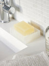 A translucent white silicone soap dish holding a pale yellow bar of soap, placed on a white bathroom sink with a tiled wall and chrome faucet in the background.