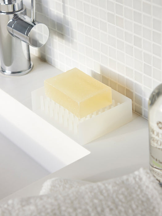 A translucent white silicone soap dish holding a pale yellow bar of soap, placed on a white bathroom sink with a tiled wall and chrome faucet in the background.