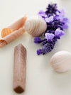 A close-up, shallow depth-of-field shot of natural stone and wood bath stones, including a lavender wood piece, a white marble sphere, and a peach-colored stone, with purple lavender flowers on a light cream background.