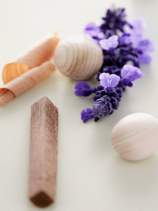 A close-up, shallow depth-of-field shot of natural stone and wood bath stones, including a lavender wood piece, a white marble sphere, and a peach-colored stone, with purple lavender flowers on a light cream background.
